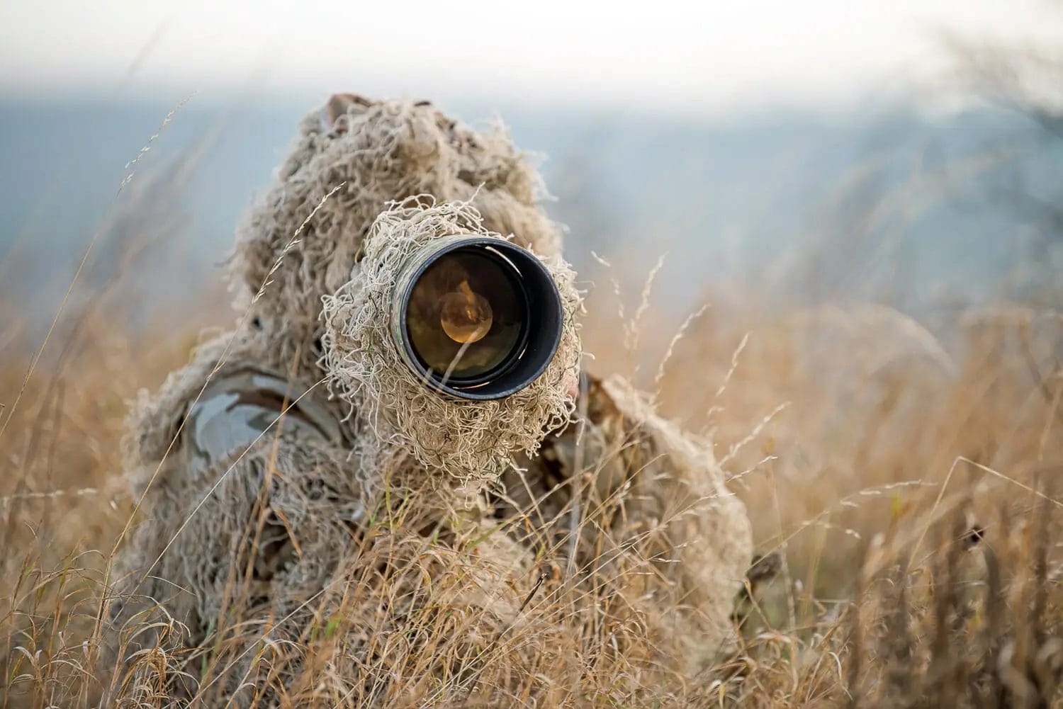 A photographer in a ghillie suit using a long lens camera in dry grassland, camouflaged to blend in with the natural surroundings.