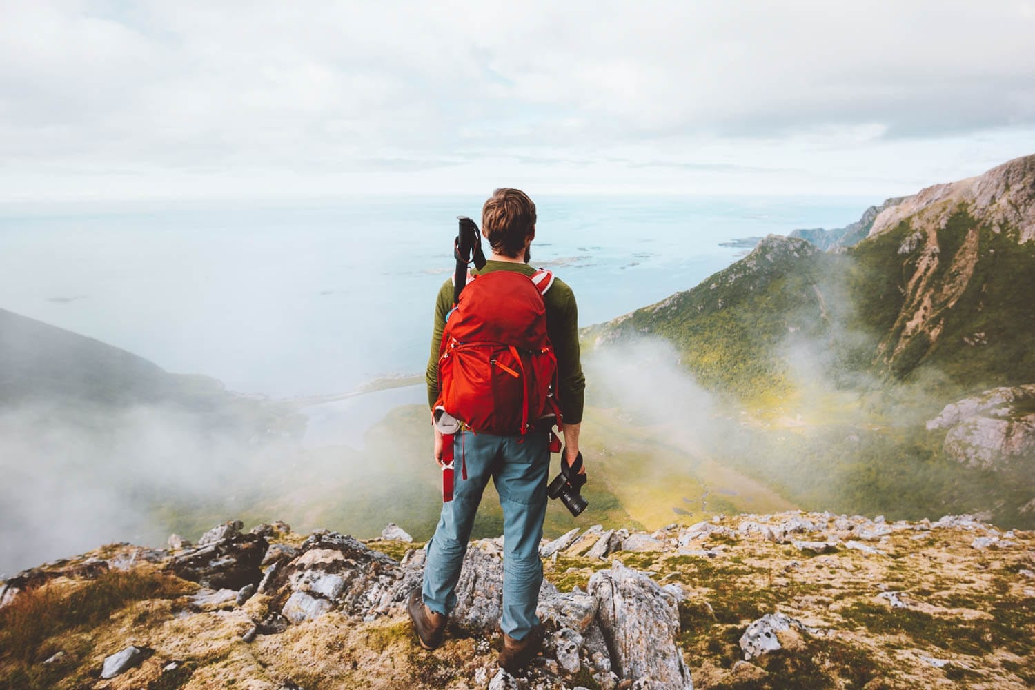 A hiker with a red backpack standing atop a mountain overlooking a misty coastline.